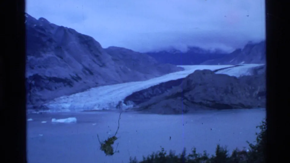 Blue-shifted Alaska glacier landscape with water in the foreground, dark hillsides, low clouds, and visible film wear.