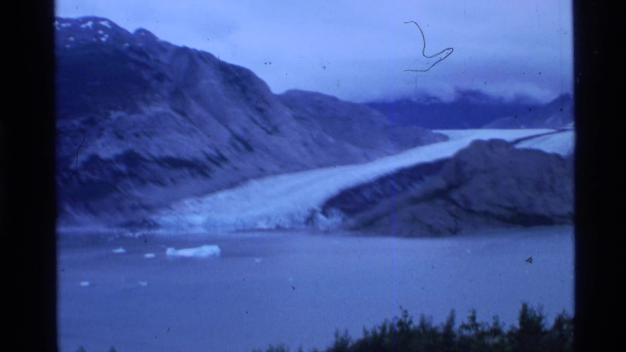 1977 8mm home-movie view of an Alaskan glacier flowing into fjord water below dark mountain slopes.