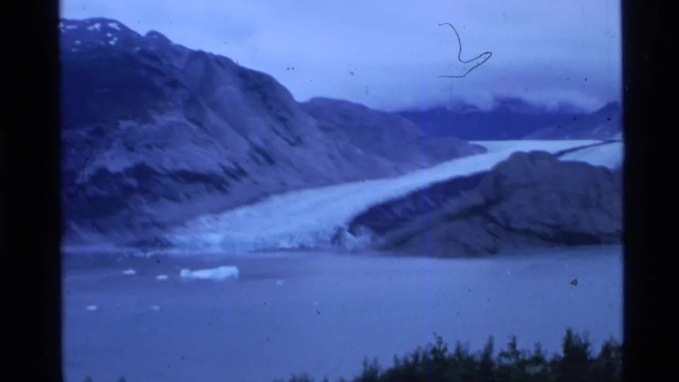 1977 8mm home-movie view of an Alaskan glacier flowing into fjord water below dark mountain slopes.
