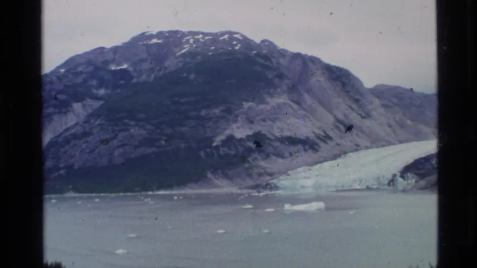 1977 Alaska 8mm frame showing a dark mountain slope, pale glacier ice, and small icebergs in fjord water.