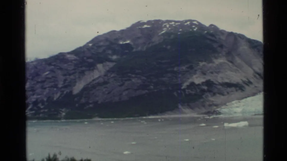 Vintage 8mm Alaska glacier view with calm fjord water, scattered icebergs, and rugged mountain terrain.