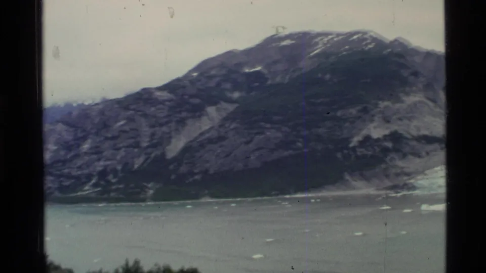 1977 Alaska 8mm frame showing a dark rocky mountain beside calm water with scattered ice and soft, faded color.
