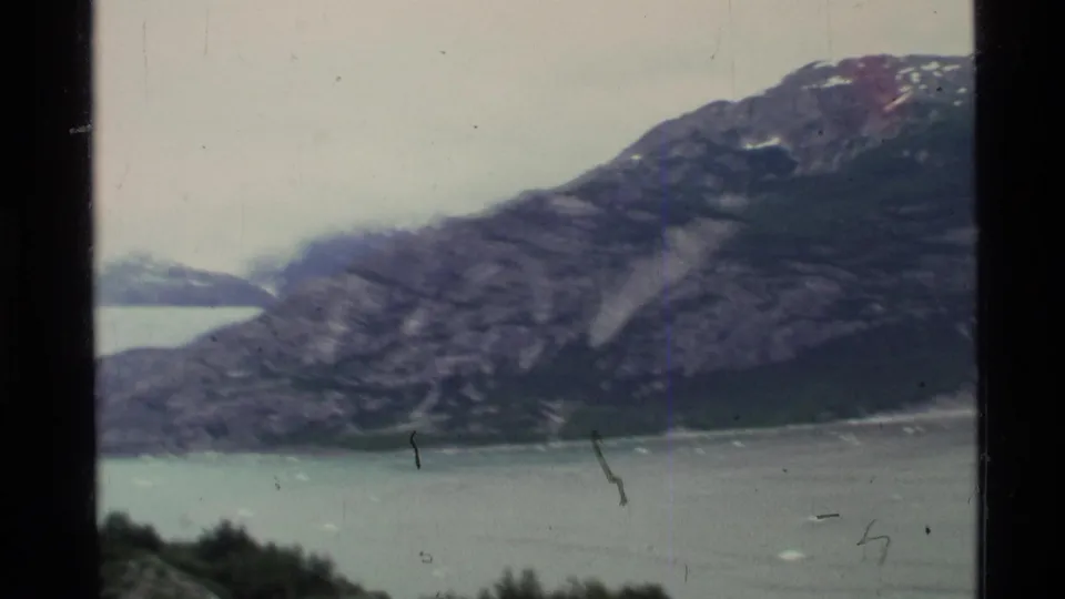 1977 Alaska 8mm frame with rocky fjord walls, distant pale glacier ice, low cloud, and small ice pieces on the water.