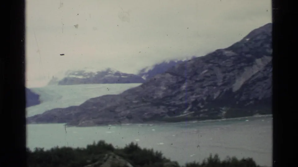 1977 Alaska 8mm frame showing a pale glacier beyond a rocky shoreline, calm fjord water, and dark foreground brush.