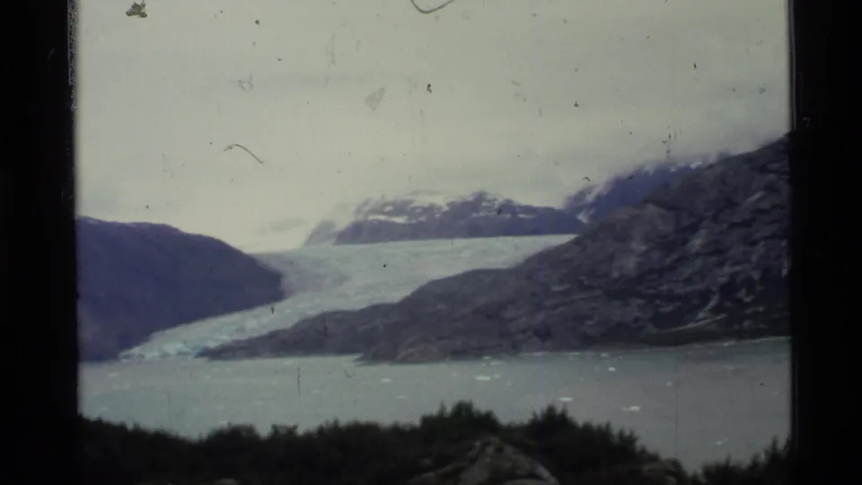 1977 Alaska 8mm frame of a broad glacier flowing between rocky mountains toward a fjord with scattered ice pieces.