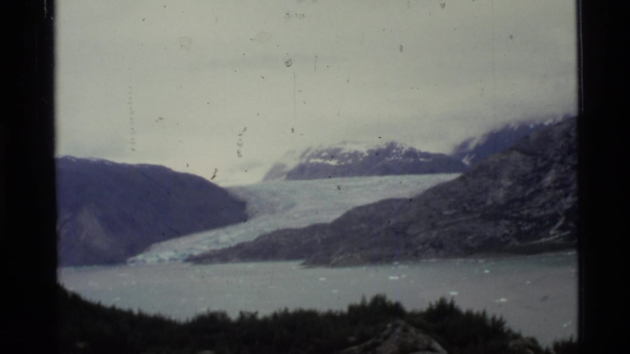 1977 Alaska 8mm frame showing a pale glacier descending into a bay, with rugged mountains and snow patches under an overcast sky.