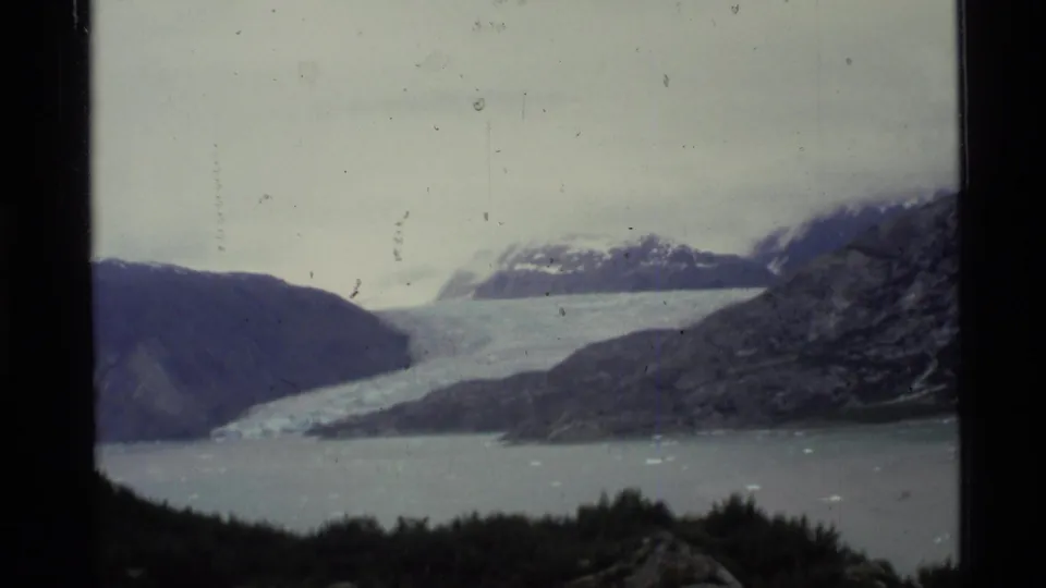 1977 Alaska 8mm frame showing a pale glacier descending into a bay, with rugged mountains and snow patches under an overcast sky.