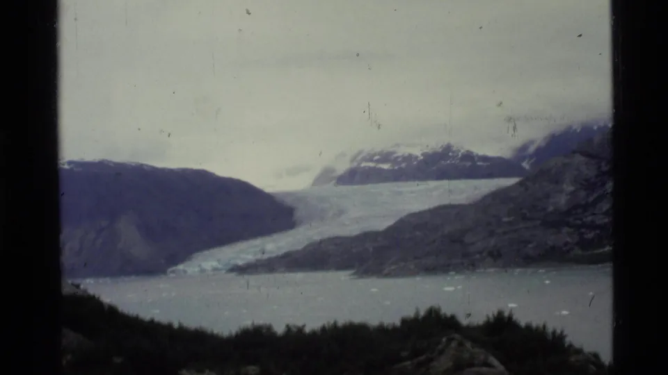 1977 Alaska home-movie frame with a distant glacier, pale water, mountain ridges, small ice pieces, and visible 8mm scan borders.