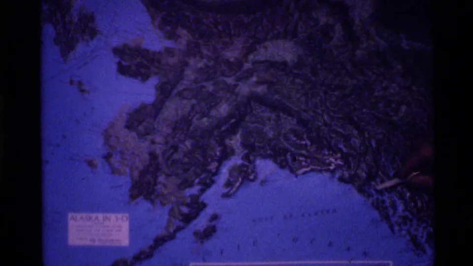 1977 Alaska clip frame showing a blue-toned relief map of Alaska with raised terrain, coastline details, and printed text near the lower left.