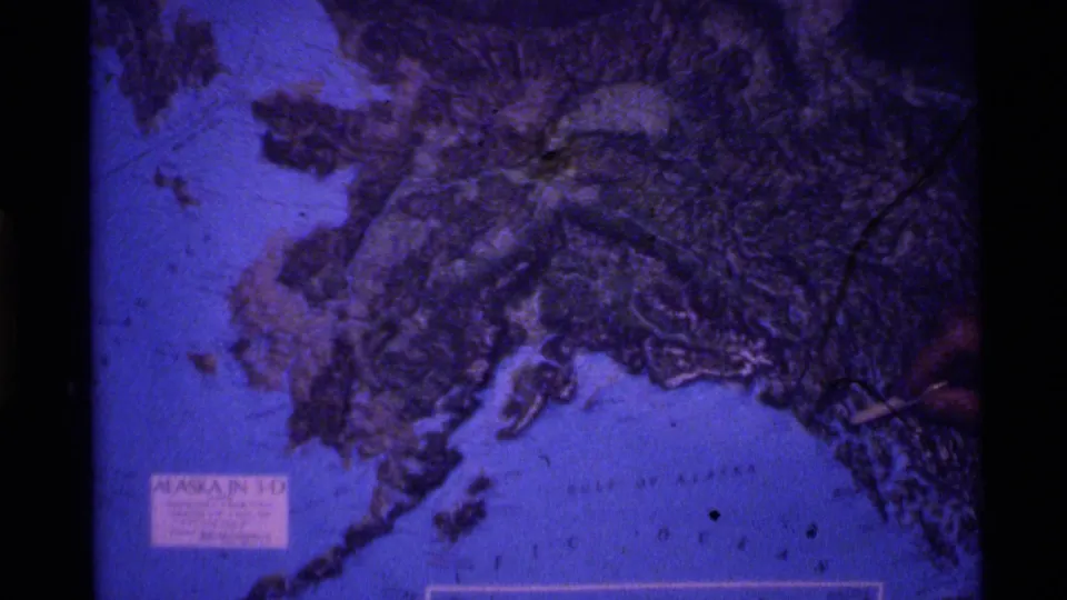 1977 Alaska home-movie frame showing a raised map of Alaska, printed labeling, and a hand pointing toward the terrain.