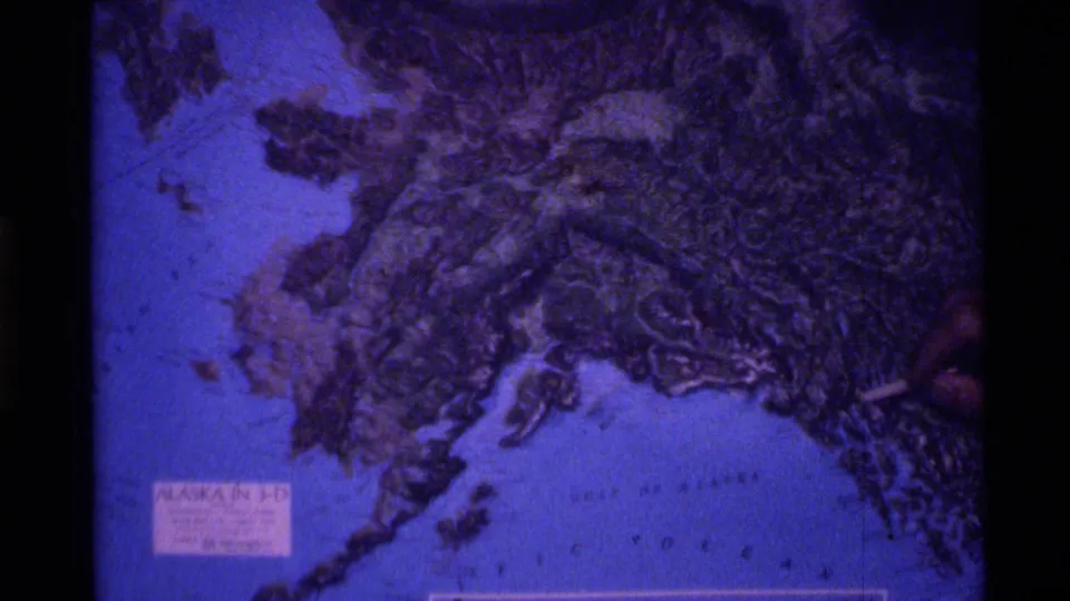 1977 Alaska home-movie frame showing the relief map again as a hand points from the right side toward the terrain.
