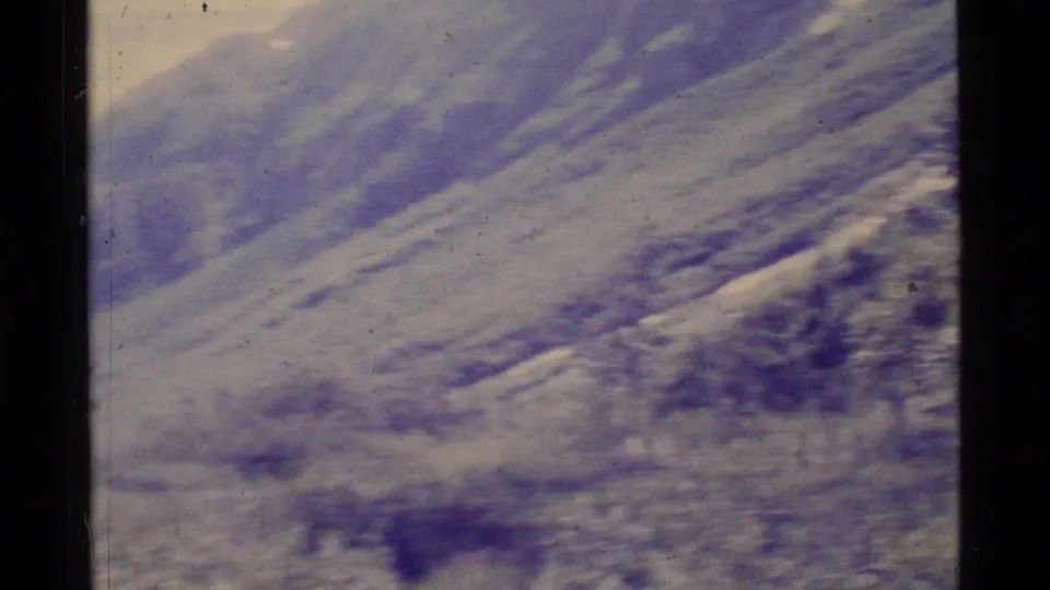 1977 8mm Alaska home-movie frame showing pale mountain slopes and a distant valley under soft, faded color.