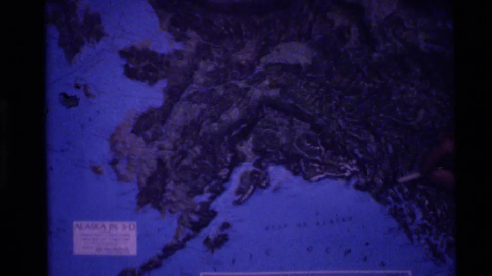 Blue-shifted 1977 home-movie frame showing a printed Alaska map used as a location reference.