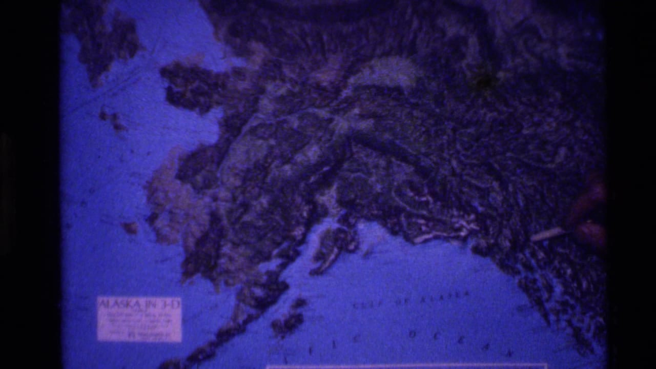 Projected relief map of Alaska with a hand-held pointer near the right edge.