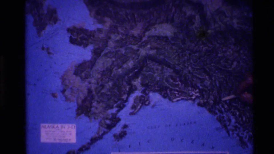 1977 8mm frame showing a projected 3-D-style map of Alaska with blue water, textured land, and a pointer at the side.