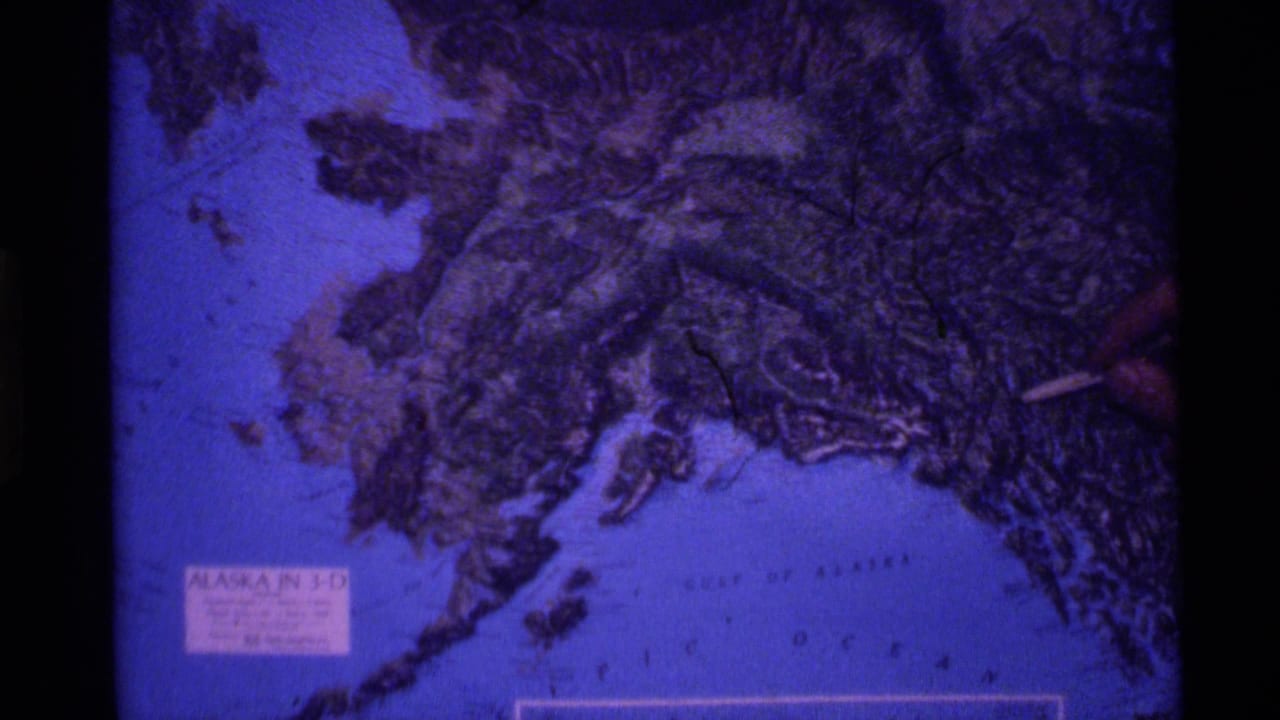 1977 8mm frame of a hand pointing across a blue-purple 3-D relief map of Alaska.