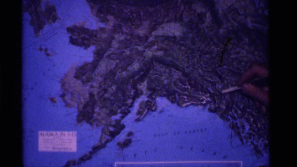A hand marks a spot on a 3-D Alaska map, with coastline and ocean labels visible in the 1977 home-movie frame.