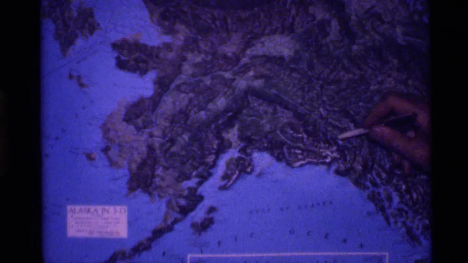 Soft 8mm frame showing a hand using a pointer on a blue-purple Alaska relief map with ocean lettering below.