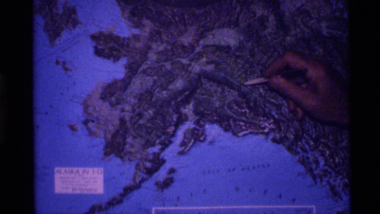 1977 8mm frame showing a hand pointing at a raised topographical map of Alaska under blue light.