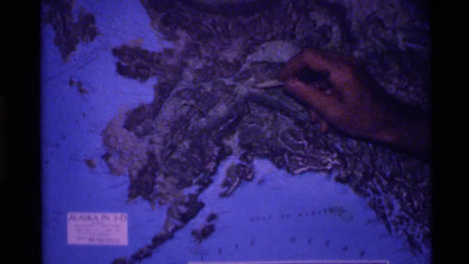 1977 home-movie frame of a pointer tracing central Alaska on a raised geography map.