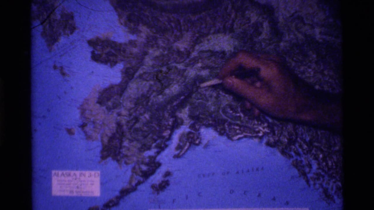 Hand holding a pointer over a printed Alaska relief map.