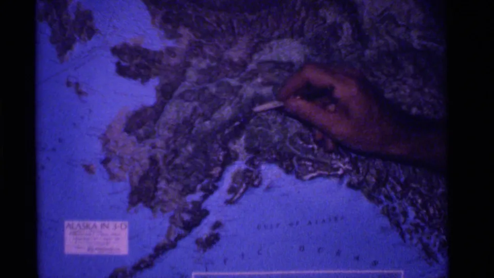 A hand marks a point on a printed Alaska terrain map in a 1970s color 8mm travel sequence.