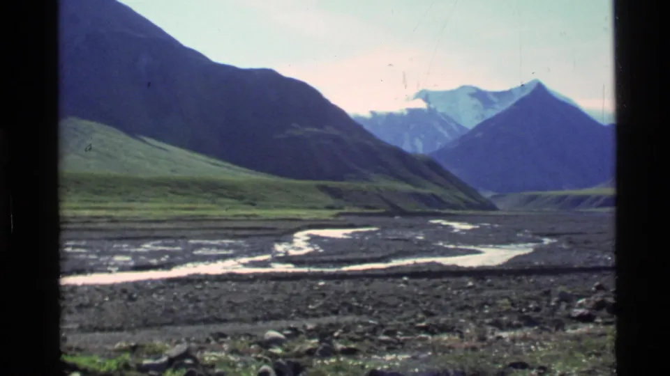 Alaska mountain valley view with green slopes, a winding river, rocky ground, and a bright sky on 8mm film.
