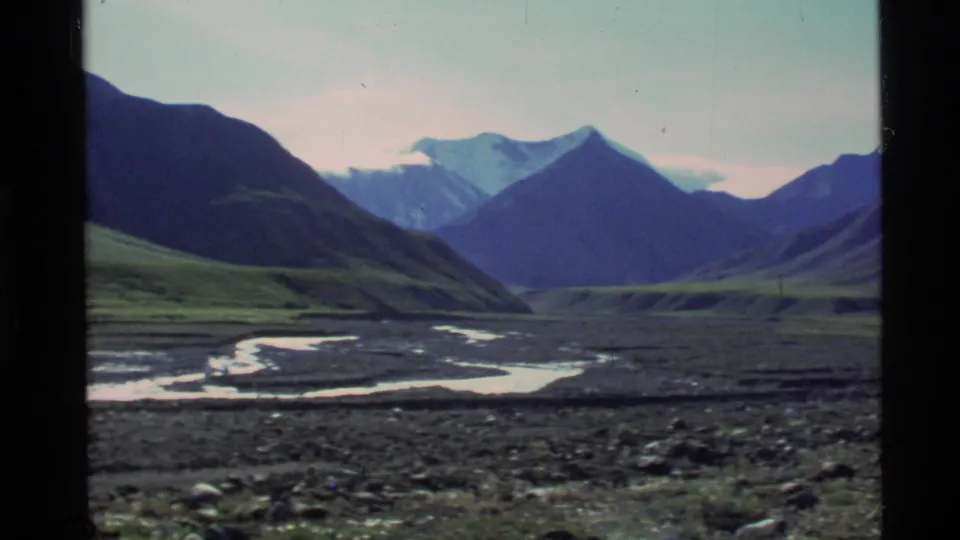 Wide Alaska landscape frame showing dark mountain slopes, a braided river course, and distant peaks under pale sky.