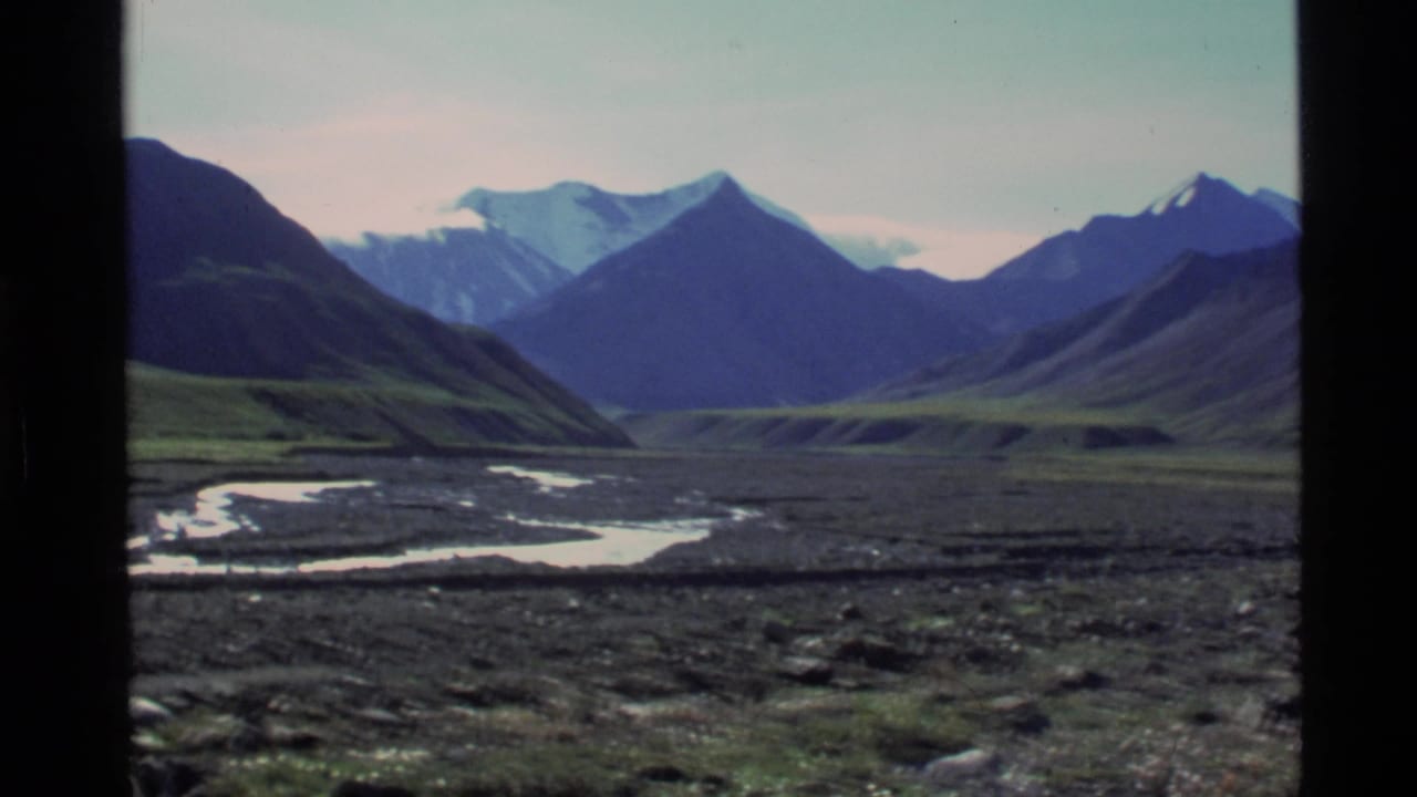 1977 Alaska 8mm frame showing a broad river valley between dark mountain slopes and distant snow-capped peaks.