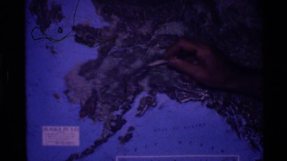 1977 Alaska 8mm frame showing a hand pointing on a blue-toned Alaska map with visible printed labels and coastal outlines.