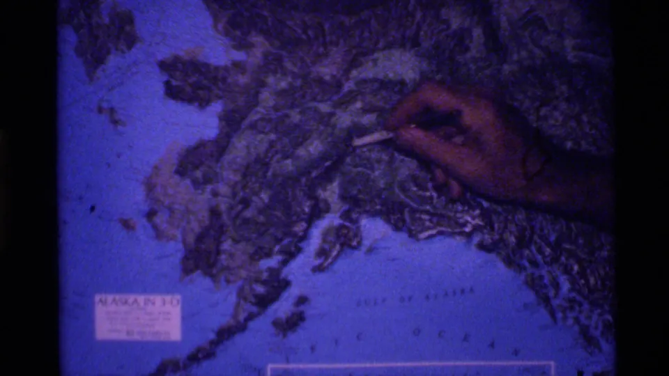 1977 Alaska 8mm frame with a hand indicating a place on a blue-toned Alaska map near visible Gulf of Alaska text.