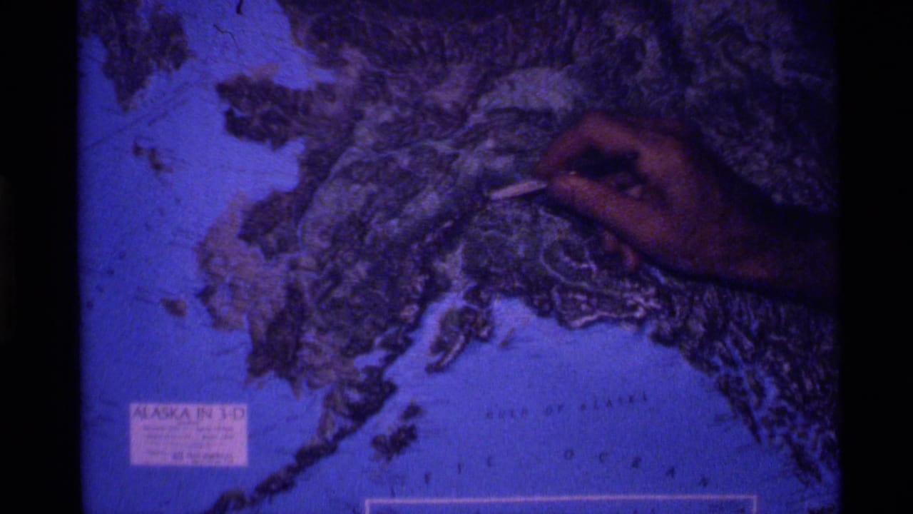 1977 8mm frame showing a hand pointing at rugged terrain on a blue-tinted Alaska relief globe.