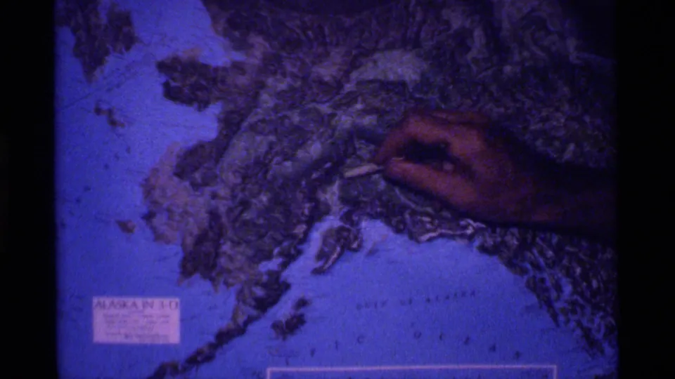 The hand and pointer move across the Alaska relief globe, with printed map labeling and heavy film grain visible.