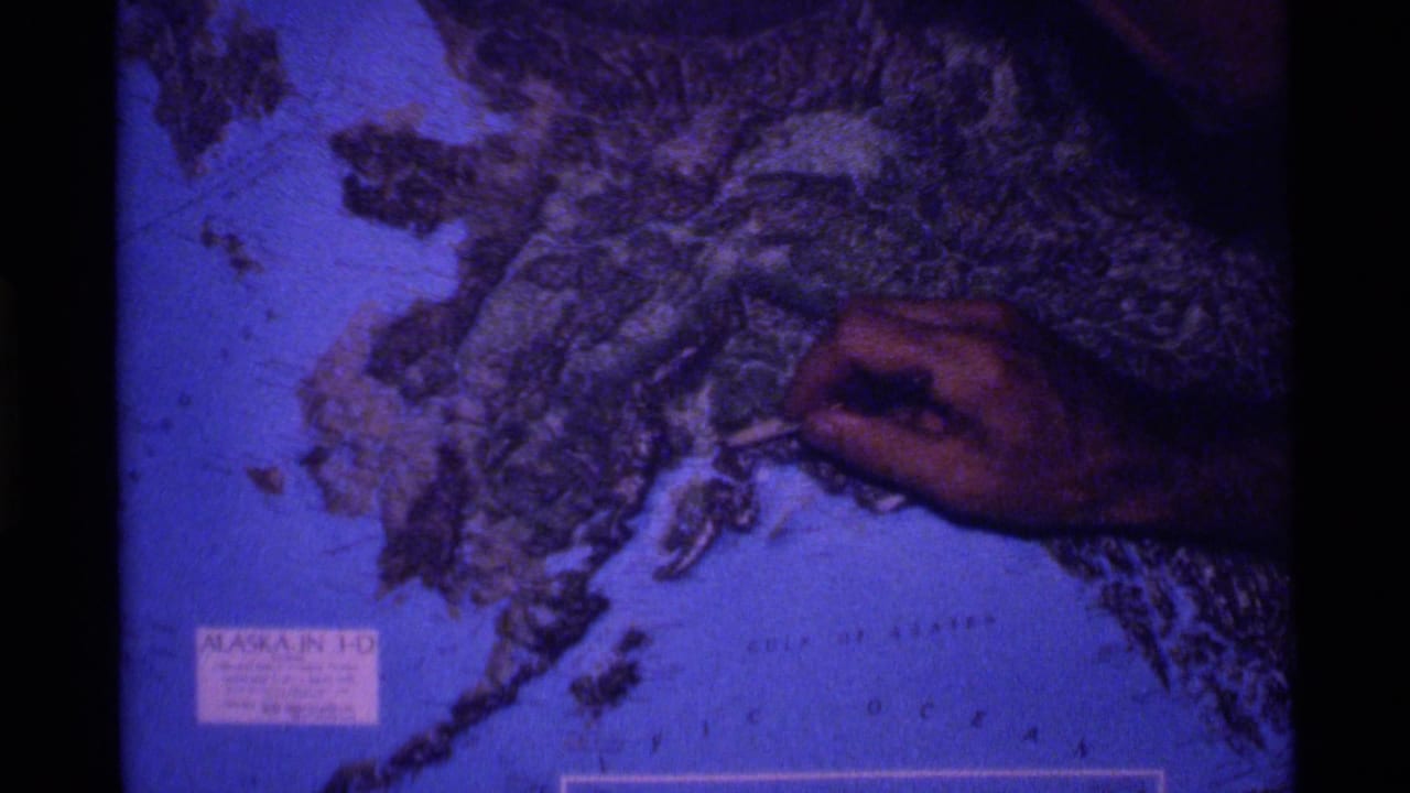 A hand points to a textured relief map of Alaska in 1977 8mm footage, with the Gulf of Alaska area and map label visible.