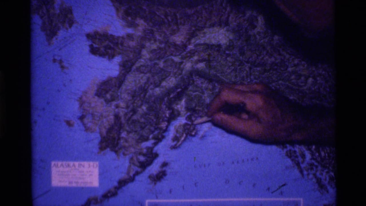 In 1977 Alaska travel footage, a hand points a pen at a raised topographical map of Alaska with visible film borders.