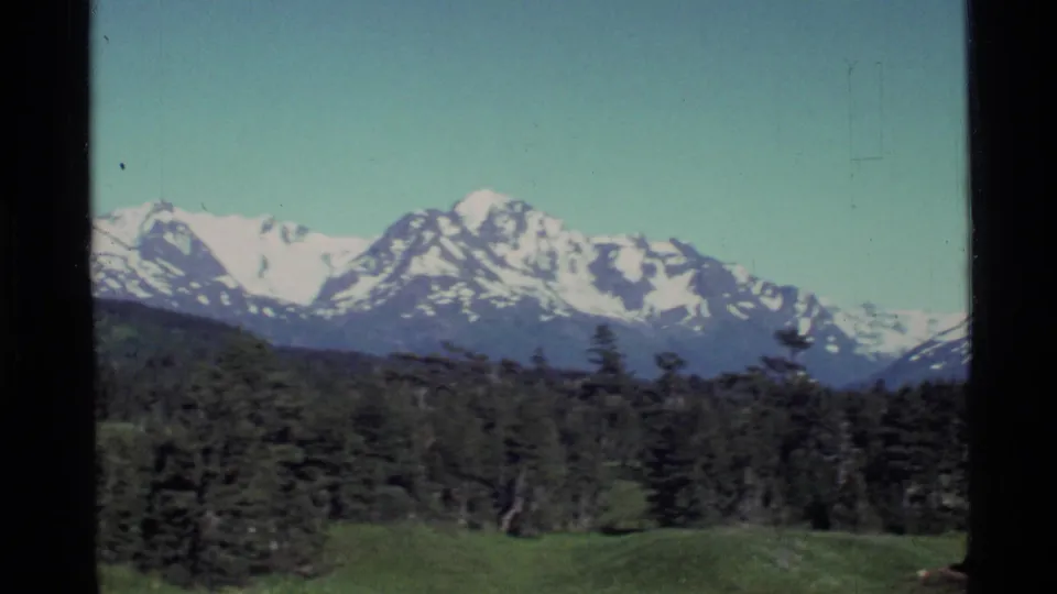 Alaska mountain peaks with patches of snow rise above evergreen trees and a grassy foreground in a vintage 8mm frame.