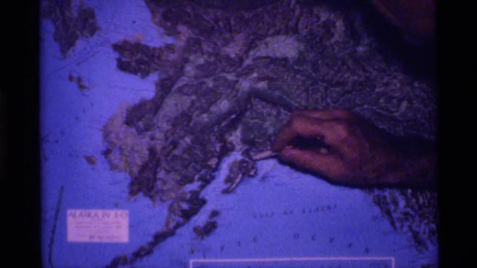 A hand indicates terrain on a relief map of Alaska, with the state label visible in the lower-left area.