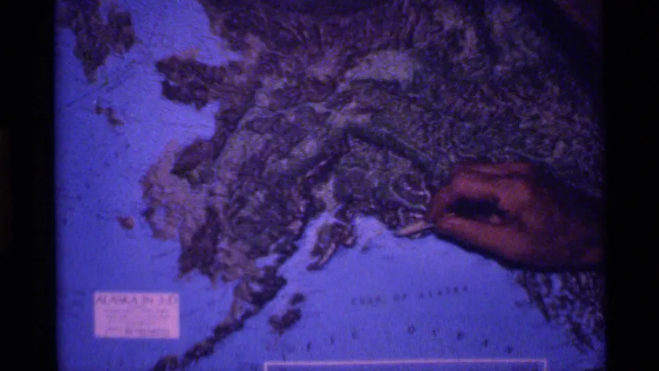 Blue-shifted 8mm frame of a hand pointing at raised terrain on an Alaska map with Gulf of Alaska lettering visible.