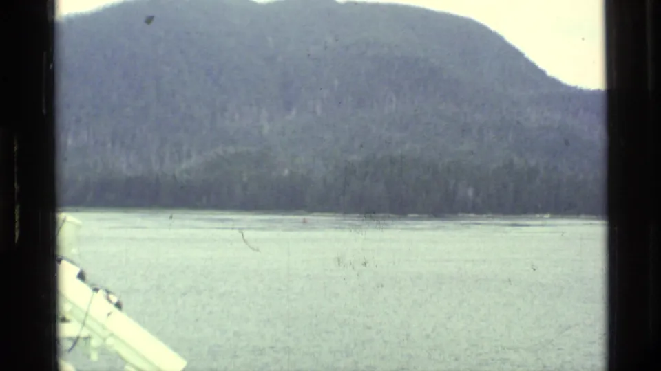 Alaska scenic view across calm water toward a forested mountain, likely filmed from a boat or shoreline in 1977 8mm footage.