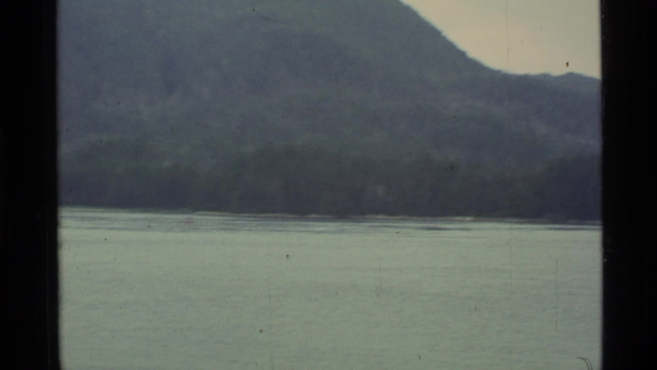 Calm lake beneath forested hills and distant mountains, framed by dark 8mm borders.