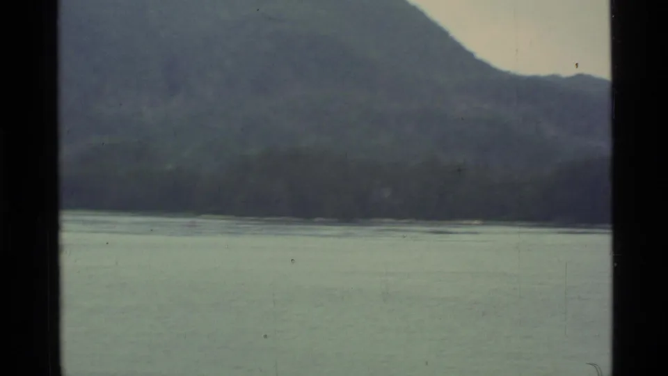 1977 Alaska 8mm frame showing a calm lake, wooded shoreline, and layered mountains under an overcast sky.