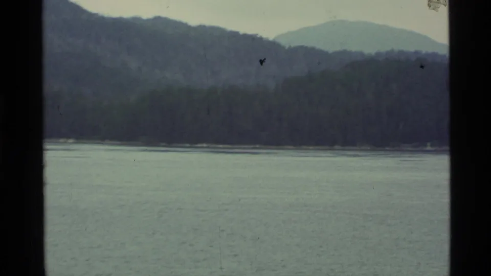 1977 Alaska home-movie view across a lake toward a forested bank and hazy mountain ridges.
