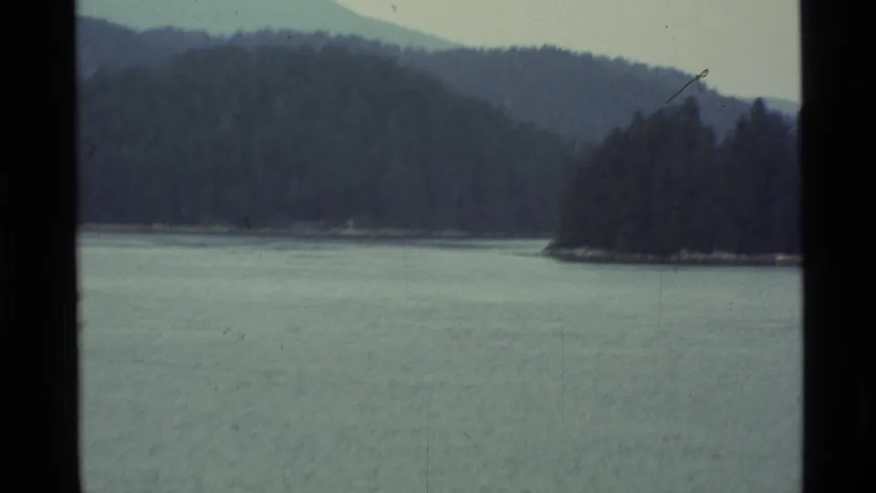 1977 Alaska 8mm frame of lake water, forested hills, and a tree-covered point along the opposite shore.