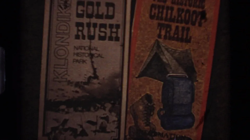 1977 Alaska 8mm frame showing printed Klondike Gold Rush National Historical Park and Chilkoot Trail materials with illustrated travel gear.
