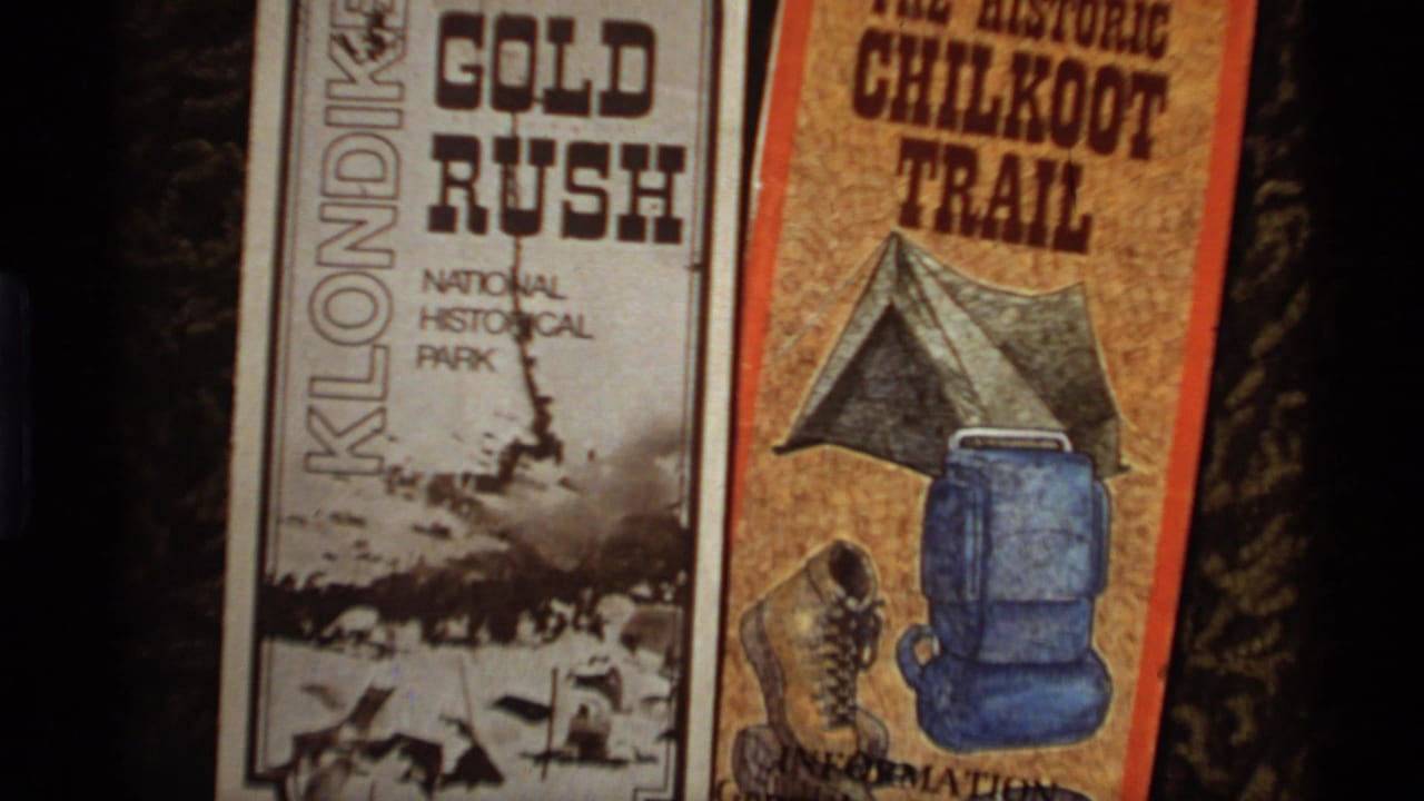 1977 Alaska home-movie frame showing Klondike Gold Rush and Chilkoot Trail brochures with miners and camping gear artwork.