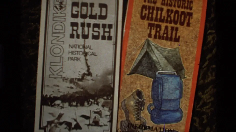 1977 Alaska home-movie frame showing printed Klondike and Chilkoot Trail visitor materials on a dark surface.