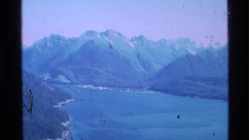 1977 Alaska 8mm aerial view of a blue fjord or inlet bordered by forested coast and snow-marked mountains.