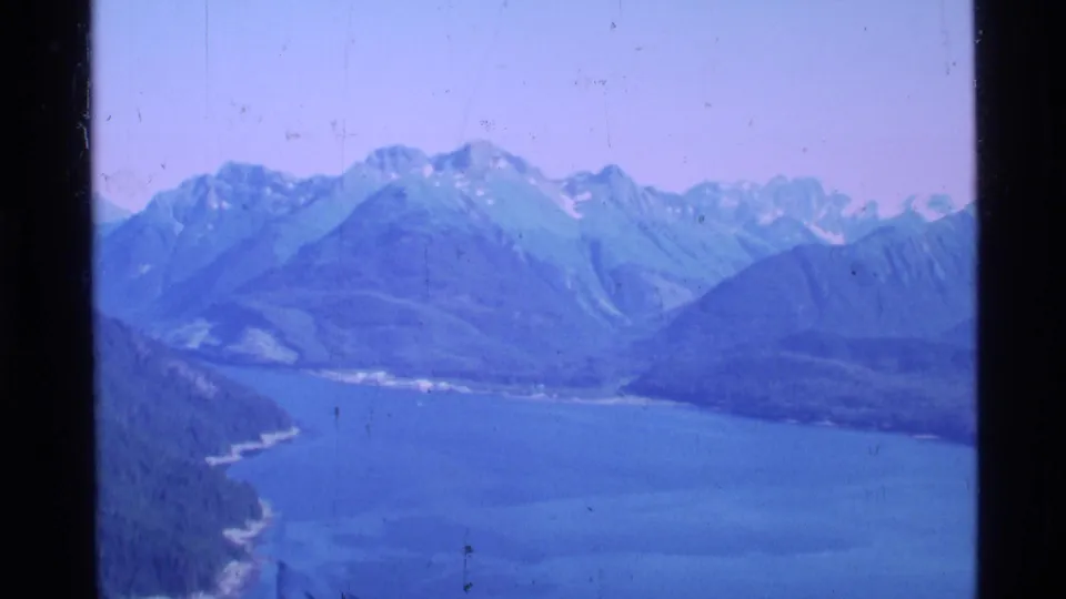 1977 Alaska 8mm frame showing an aerial view over a broad blue inlet toward rugged mountains and wooded coastline.