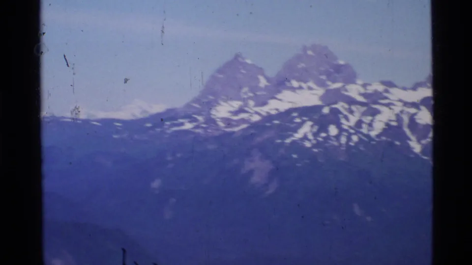 1977 Alaska aerial home-movie frame showing rugged snow-streaked peaks under a pale blue sky.