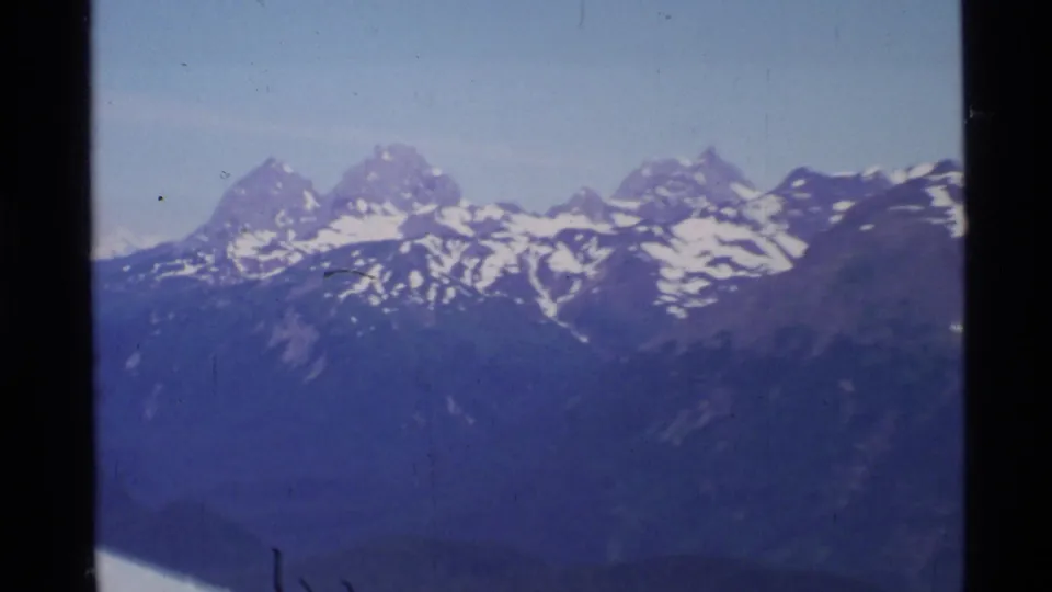 Wide 8mm aerial view of Alaskan snowy mountain ridges, partly framed by the aircraft wing below.